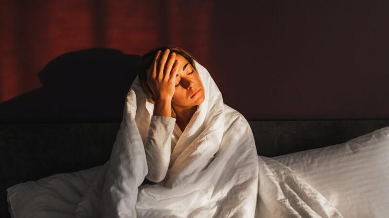A female sits on a bed, wrapped up in a duvet, holding her hand to her forehead with her eyes closed.