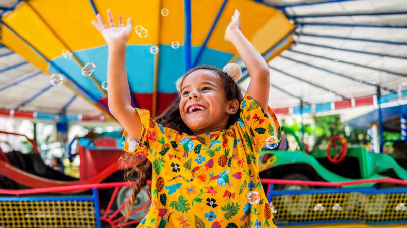 A girl wearing a bright yellow top raises her hands in the air and smiles, standing in front of a fairground ride.