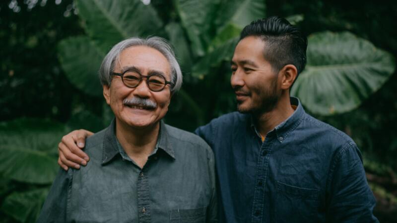 Adult son puts arm around elderly father's shoulder, standing in front of plants