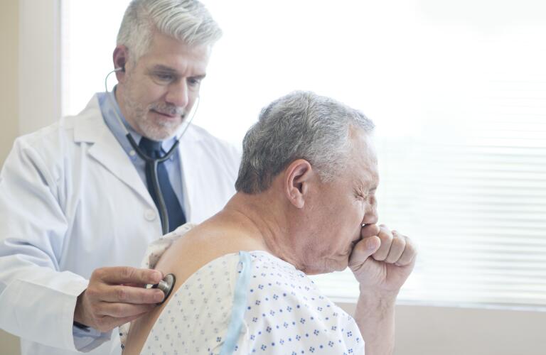 Senior Caucasian man coughing during doctor's exam while doctor checks back with stethoscope