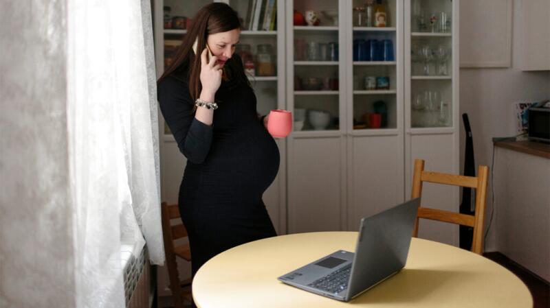 Pregnant female standing and talking on phone while looking at laptop on table