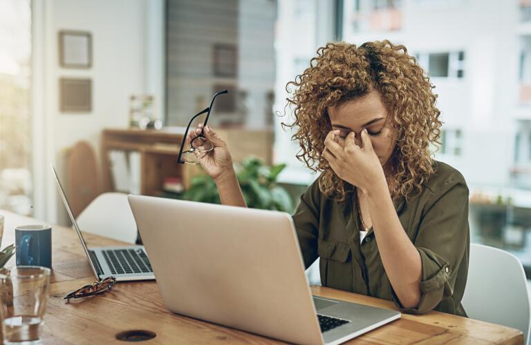 stressed young woman removing glasses and rubbing eyes in office in front of laptop