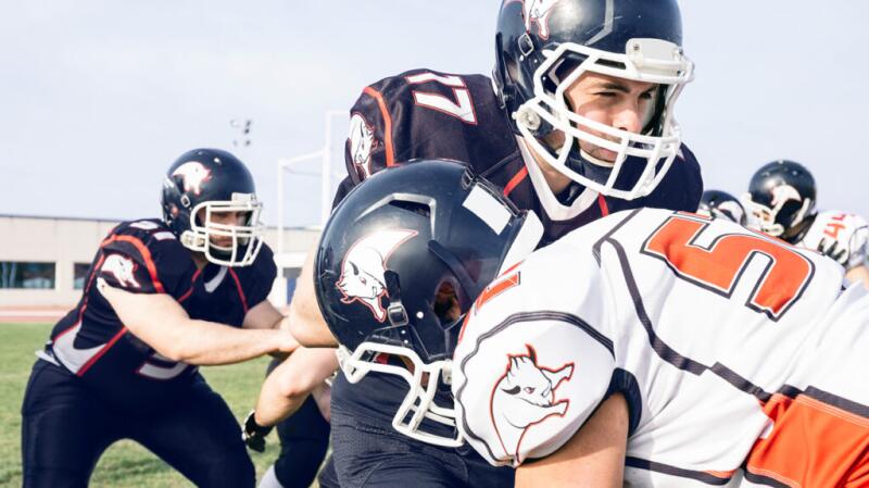 Three American football players tackle each other on an outdoor pitch, wearing blue and white uniforms and helmets.