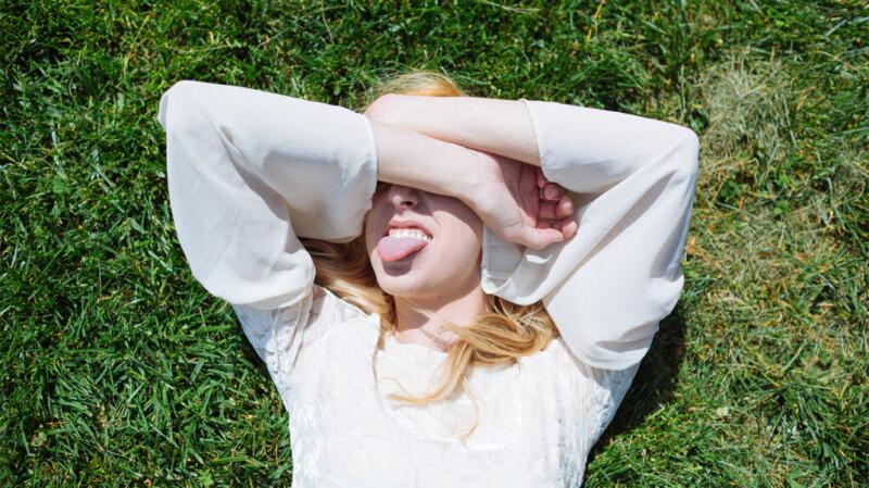 Young woman lying in grass with arms over eyes and sticking out tongue