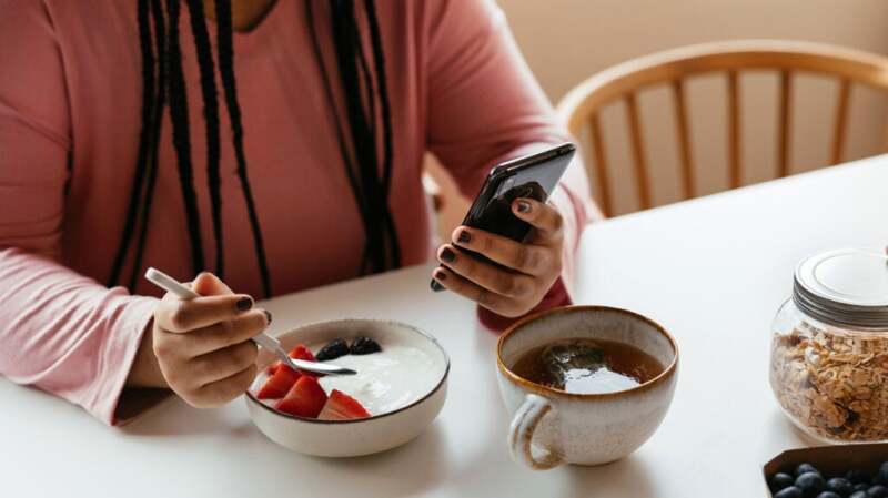 Female sitting at a table eating yogurt and looking at her phone