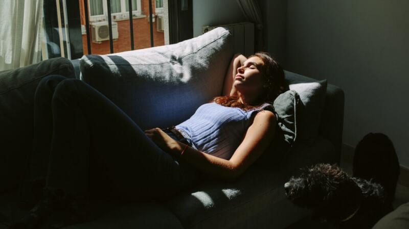 A woman is lying on a couch beside a window.