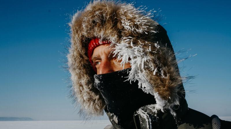 A person wears a hooded coat and face covering covered in frozen icicles.