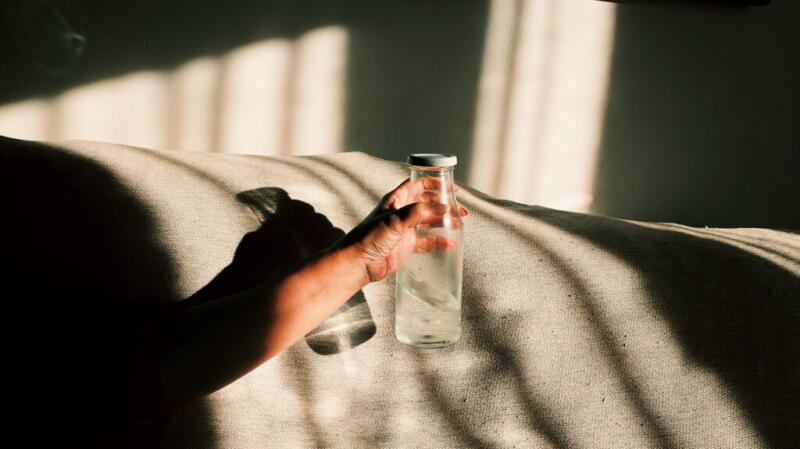 Unseen person's hand holding glass bottle of water in sunlit room
