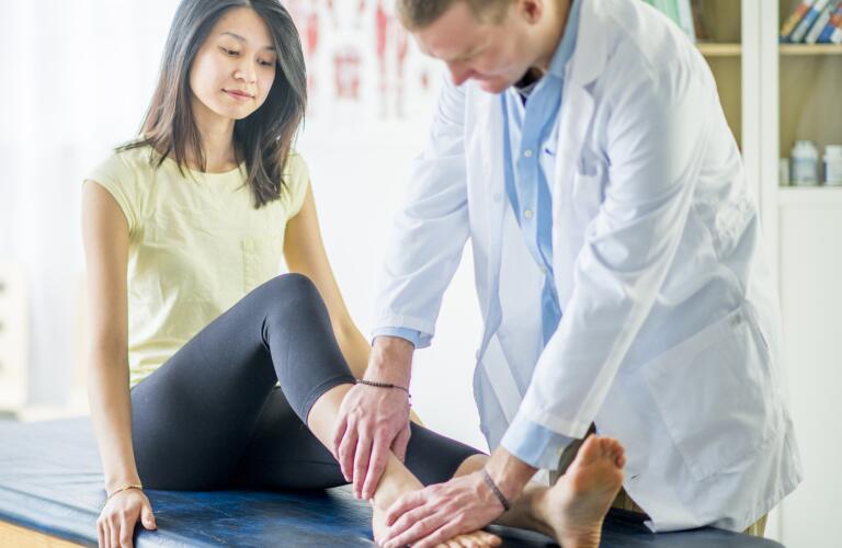 Young Asian American female patient getting ankle and foot examined by male Caucasian doctor
