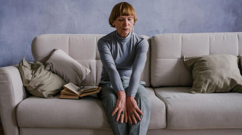 Older woman dressed in gray sitting on couch with hands on legs and eyes closed