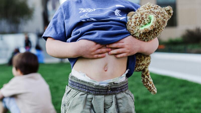 A child places their hands to their stomach while holding a stuffed toy.