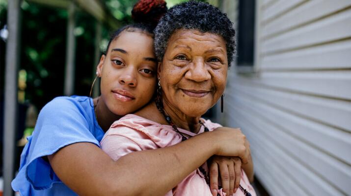 Teenage girl embracing grandmother from behind with both looking to camera with slight smiles