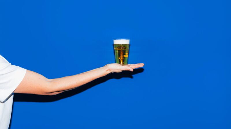 Unseen person's arm holding very small beer in palm of hand against bright blue background