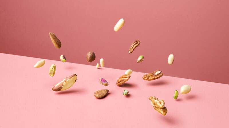 Brazil nuts and other nuts fall onto a plain pink background. 