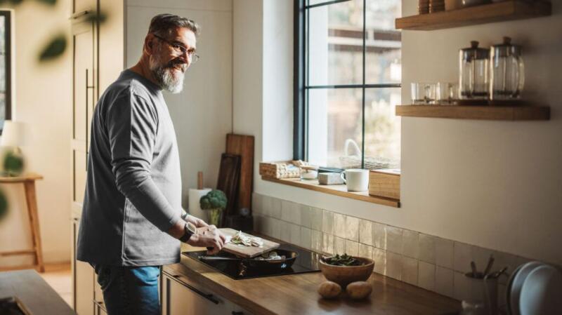 An older man smiles as he puts food into a pan to cook.