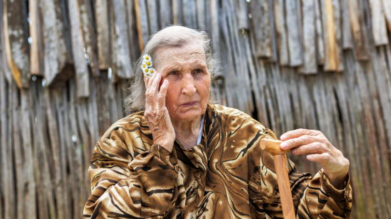 Elderly woman sits and touches hand to the side of her face with concerned expression.