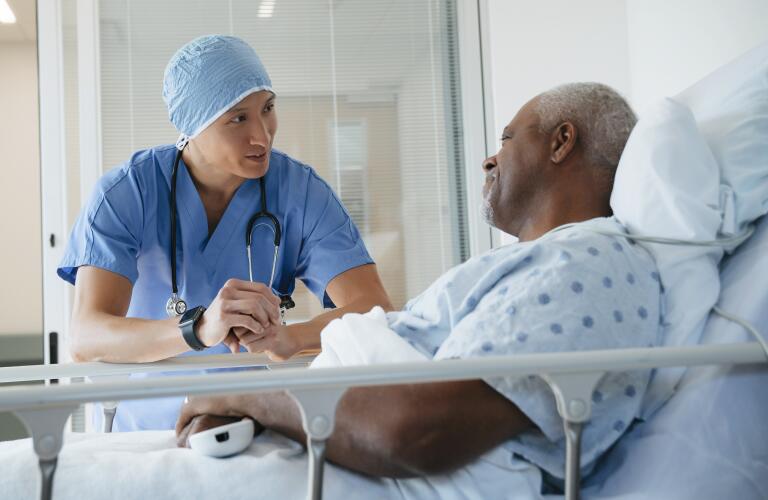 Older African American male patient in hospital bed talking to Asian American male surgeon