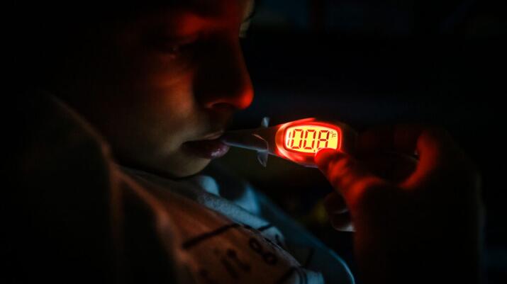 A child in a darkened room has their temperature taken with an oral thermometer. The thermometer lights up to show a reading of 100.8°F.