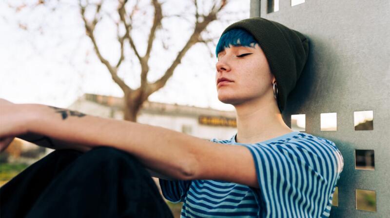 A young person is leaning against a metal post with their eyes closed.