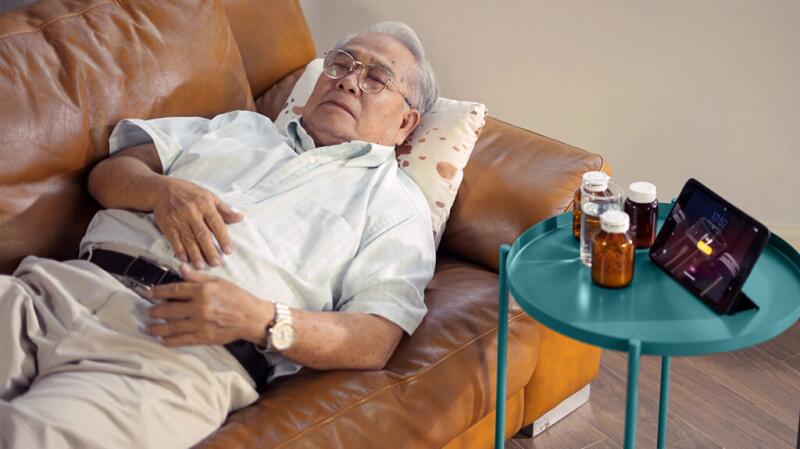Senior man lying on couch with hand over stomach and medication bottles on nearby table