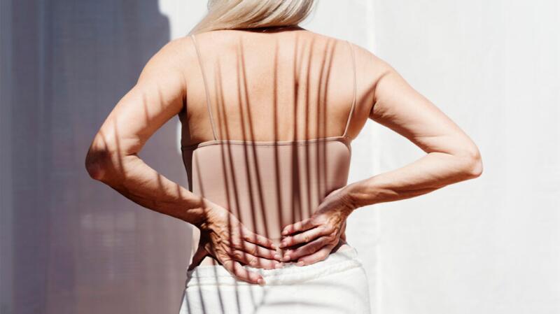 Photo from behind older female holding hands on back wearing skin-colored tank top in sunny weather