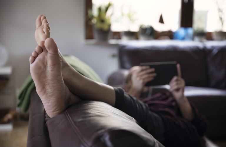 man relaxing on sofa in living room with feet up and reading on a handheld device 