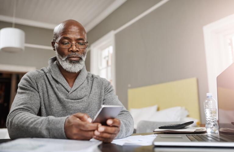 senior African American man holding smartphone in front of laptop