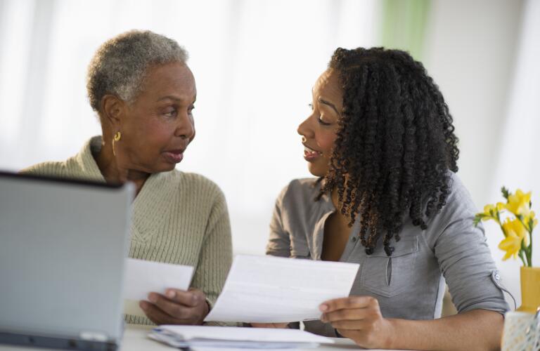 Senior African American woman talking to adult daughter about paperwork or bills at laptop