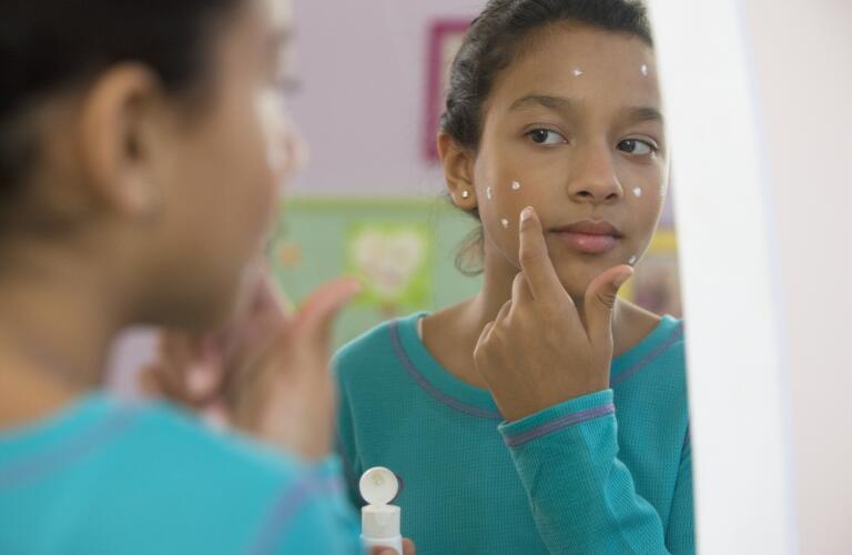Young Hispanic girl applying acne cream to spots on face in mirror