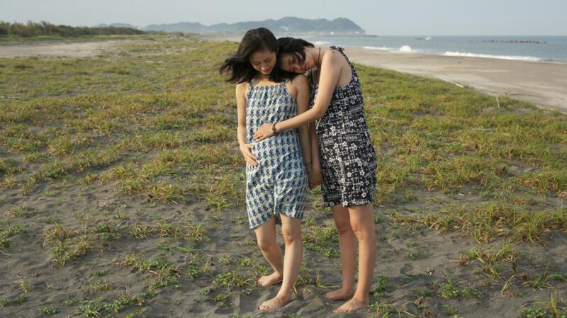 Pregnant couple standing on grass near beach with one woman holding arm across belly of other woman