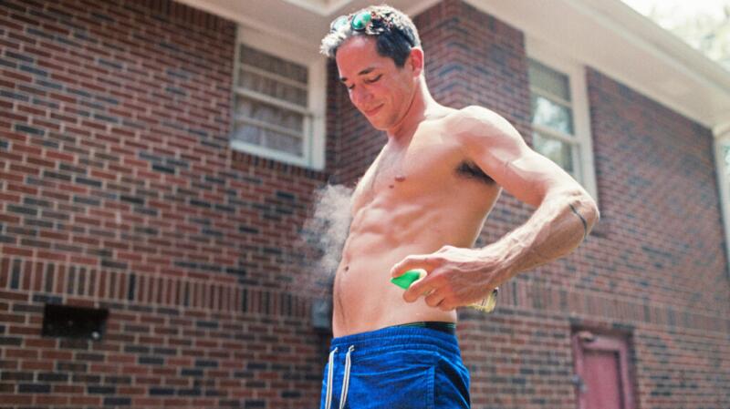 An adult male stands outside in shorts with no top, spraying aerosol insect repellant over his bare skin.