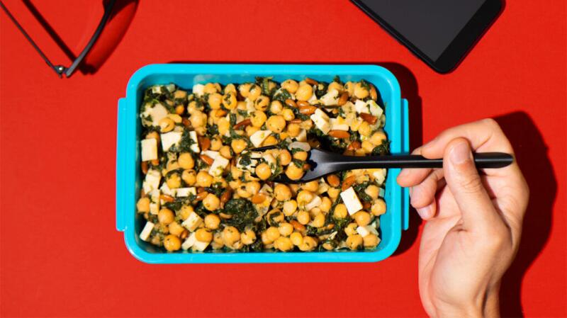 An aerial view of a blue container of chickpea salad that sits on a solid red surface. A hand holding a fork enters the frame.