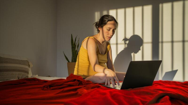 mujer embarazada en su cama usando una computadora portátil con sombras en la pared de la luz del sol entrante