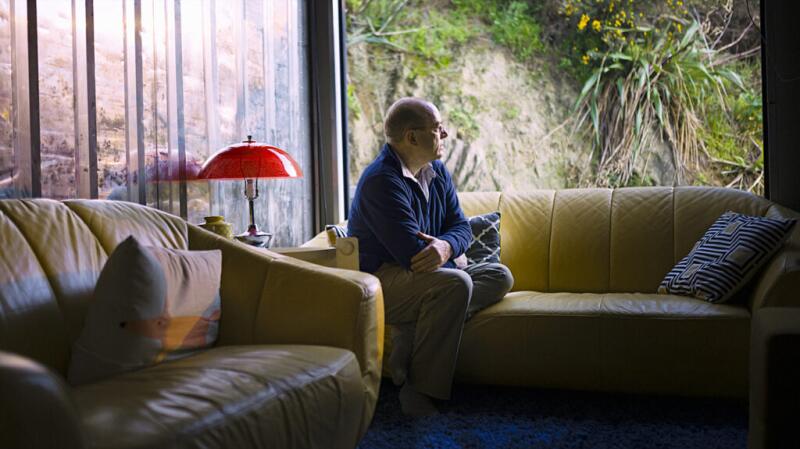 Older man sitting on a couch looking outside with a pensive expression