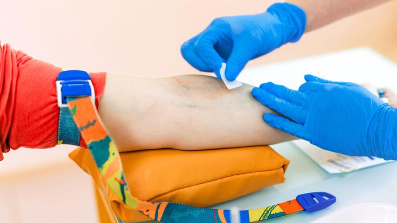 A person's arm rests on a clinician's table with a tourniquet around the upper arm. Gloved hands wipe the inner arm with a sterile wipe.