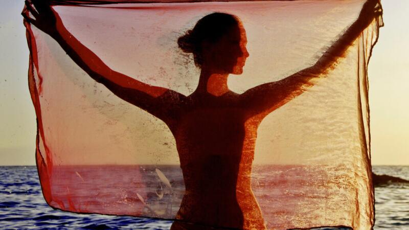 Female holding a scarf behind her on a beach, facing the water