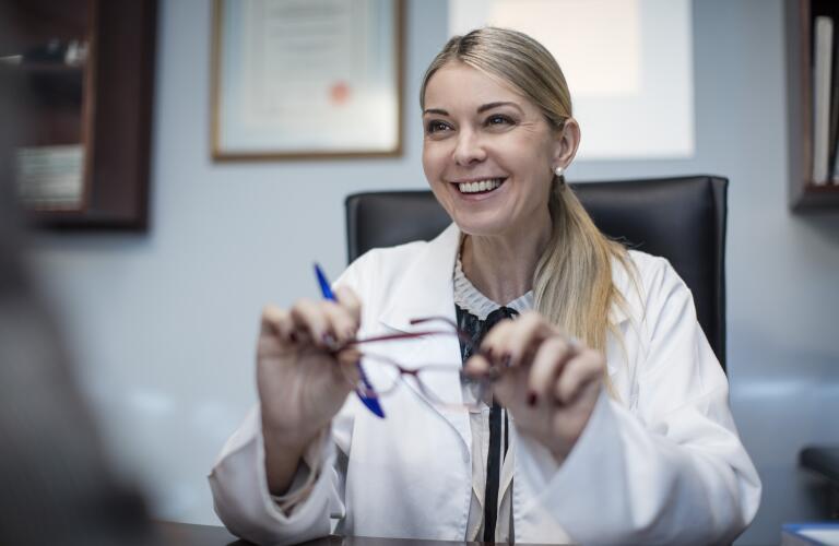 portrait of smiling Caucasian female eye doctor at desk holding eye glasses and pen