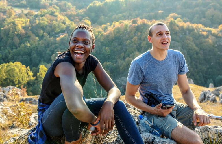 young adult black male and young adult white male with a prosthetic leg smiling and sitting on a rock in the mountains, enjoying the vista