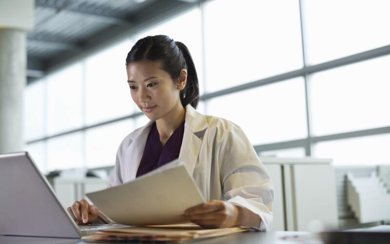 Young Asian American female doctor looking at laptop holding papers