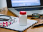 bottle of medicine and blister pack of pink and white capsules, on doctor's desk with stethoscope, eye glasses and computer in background