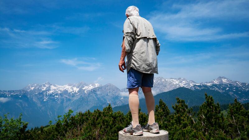 Older man wearing compression socks standing on mountain peak against bright blue sky