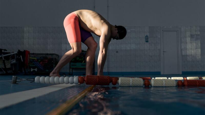 Swimmer stands at the edge of an indoor pool, bending down ready to dive in.