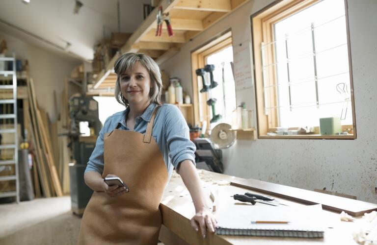 Young Caucasian woman in woodshop wearing work apron holding smartphone