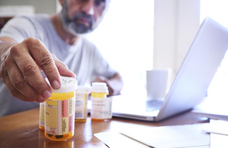 HIspanic man in his 50s reaches for one of his medicine bottles while sitting at table in front of laptop computer