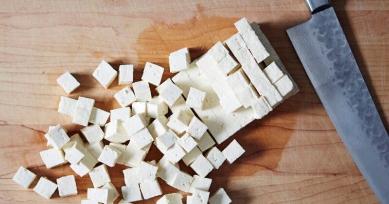 A block of tofu rests on a cutting board next to a knife.