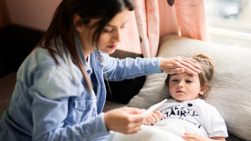 An adult feels the forehead of a toddler and reads a thermometer's temperature.