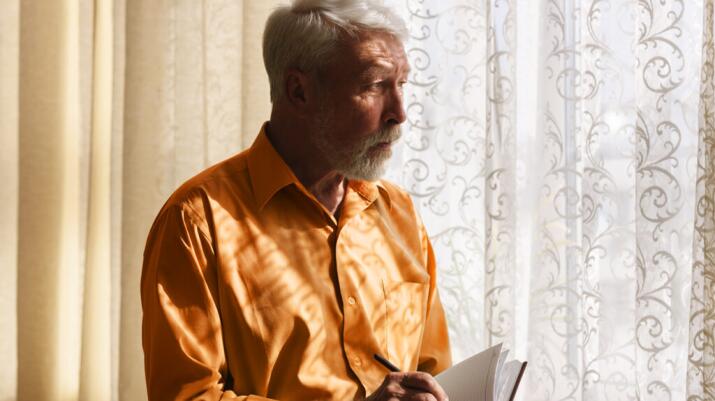Older man writing in notebook and looking out window pensively