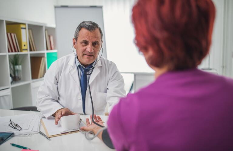 Rear view of red-headed woman getting blood pressure check from older Caucasian male doctor at desk