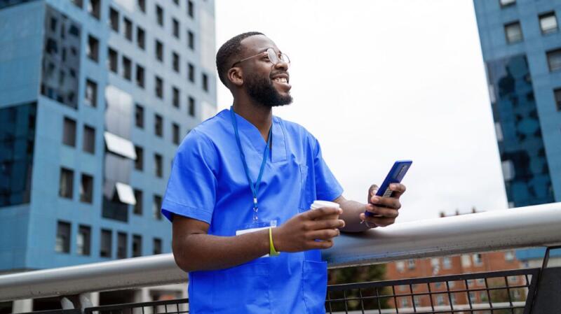 A healthcare professional standing outside, holding a cup of coffee and a smartphone.