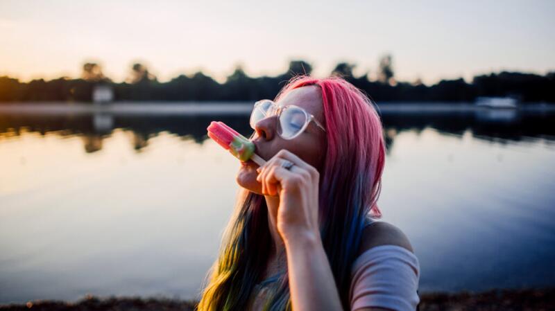 Woman with pink hair eating pink ice pop outside near lake